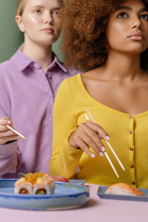 Free Two women savor sushi with chopsticks at a vibrant dining table. Indoors, lively atmosphere. Stock Photo