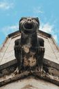 From below of shabby stone statue of gargoyle on wall of aged construction under blue sky
