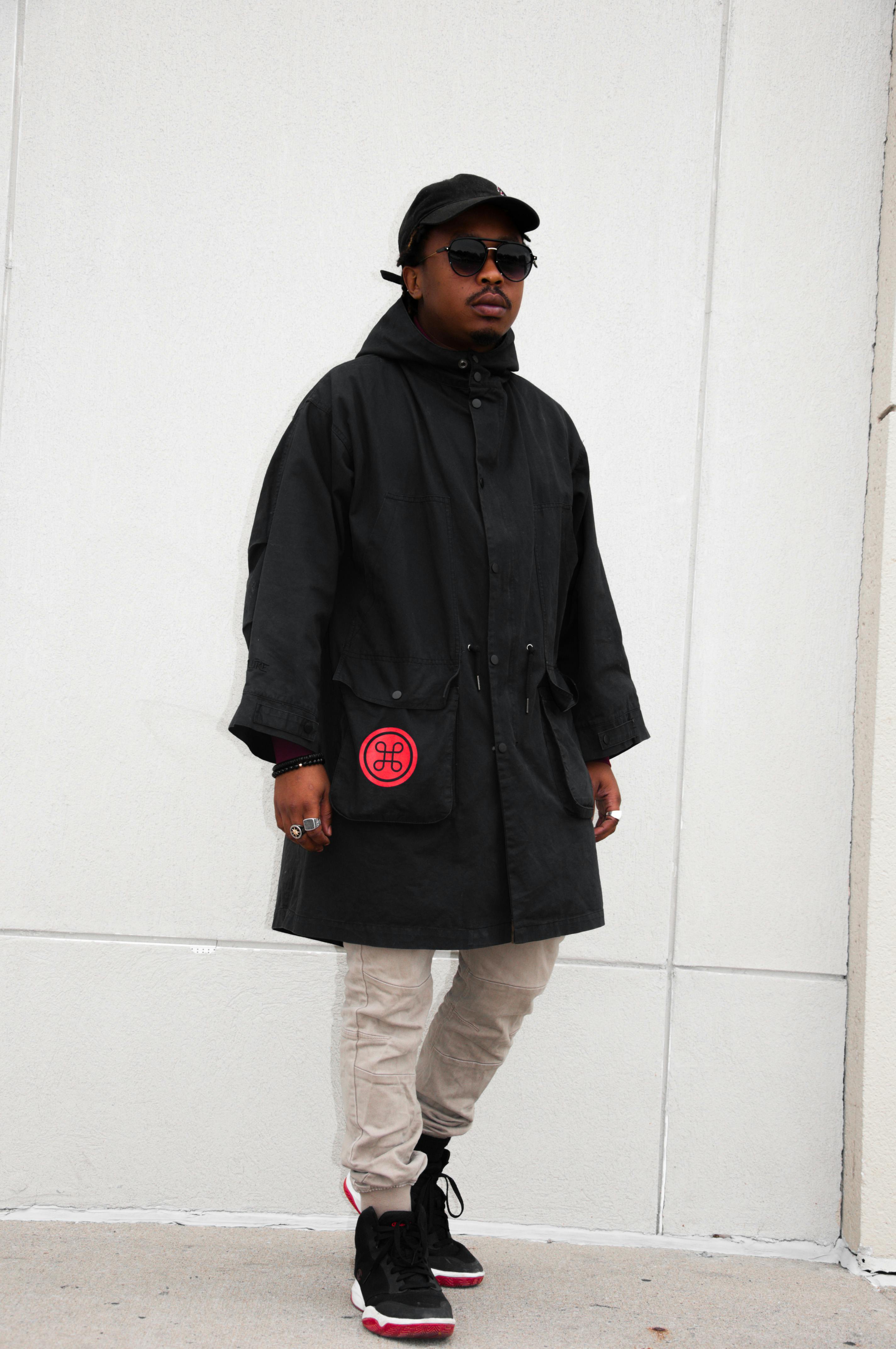 Fashionable adult in urban streetwear with black coat and sunglasses, striking a pose.