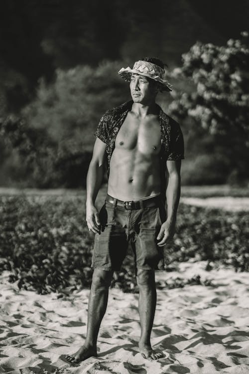 Black and white of muscular man with naked torso in straw hat looking away and standing on sand near plants