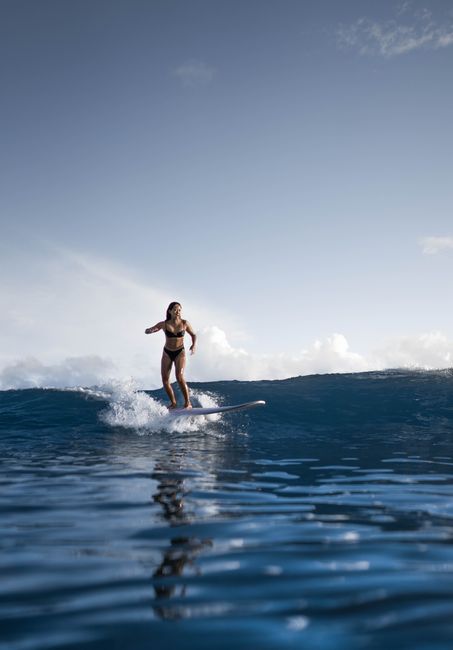 Faceless sportswoman in black swimwear riding wave while balancing on surfboard on background of cloudy blue sky in sunlight