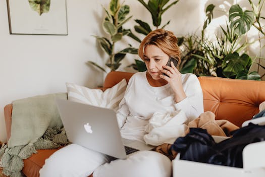 A woman works remotely on a laptop and phone, surrounded by potted plants.