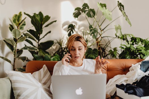 Woman multitasking with laptop and phone on a cozy sofa surrounded by lush indoor plants.