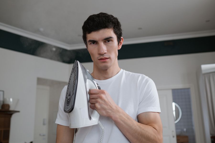 Young adult man holding a steam iron in a contemporary living room, preparing for housework.