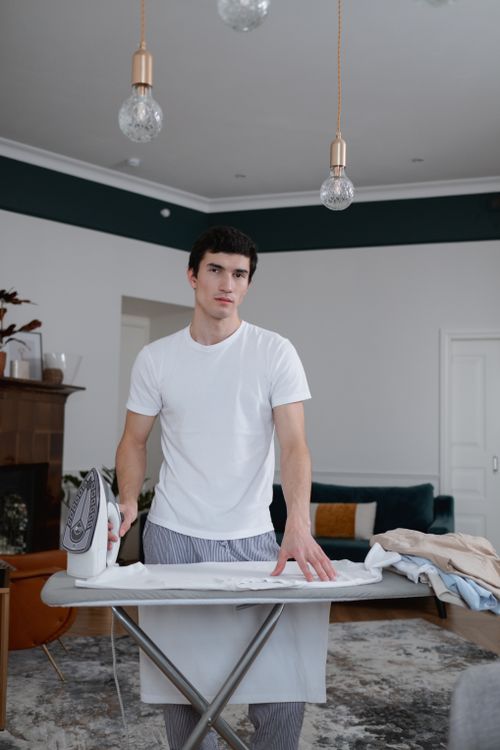 Free Young man ironing clothes in a stylish living room, showcasing a modern and tidy home setting. Stock Photo
