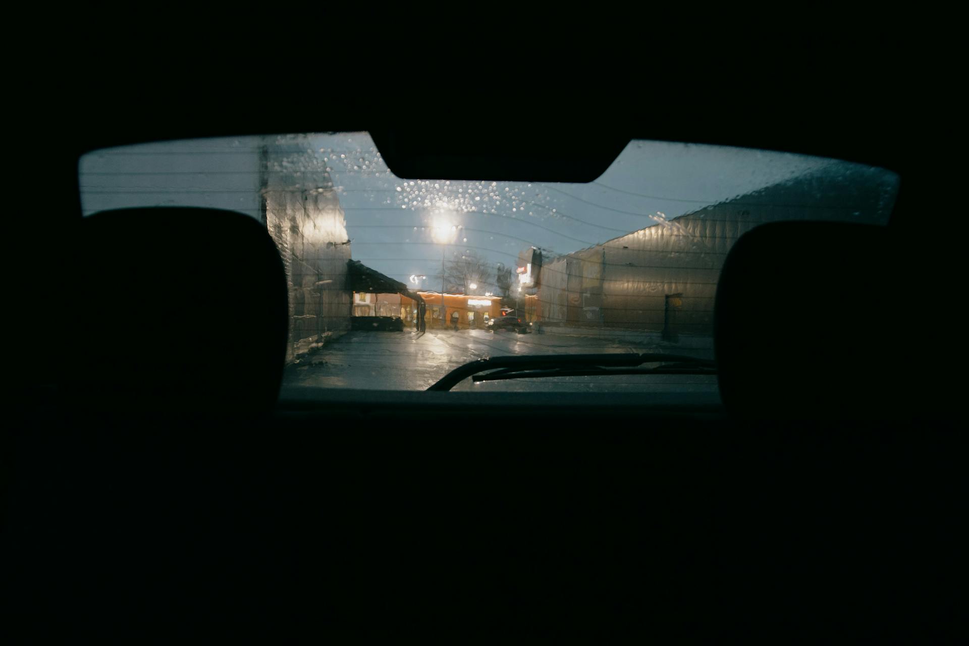 View of rainy street with buildings through wet rear window with wipers of dark automobile in city on evening time