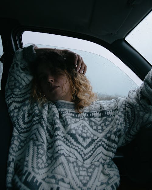 Calm female wearing warm sweater sitting in driver seat with hand on head in automobile with opened car in rainy weather