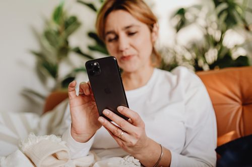 Free Woman comfortably sitting indoors using a smartphone with plants in the background. Stock Photo
