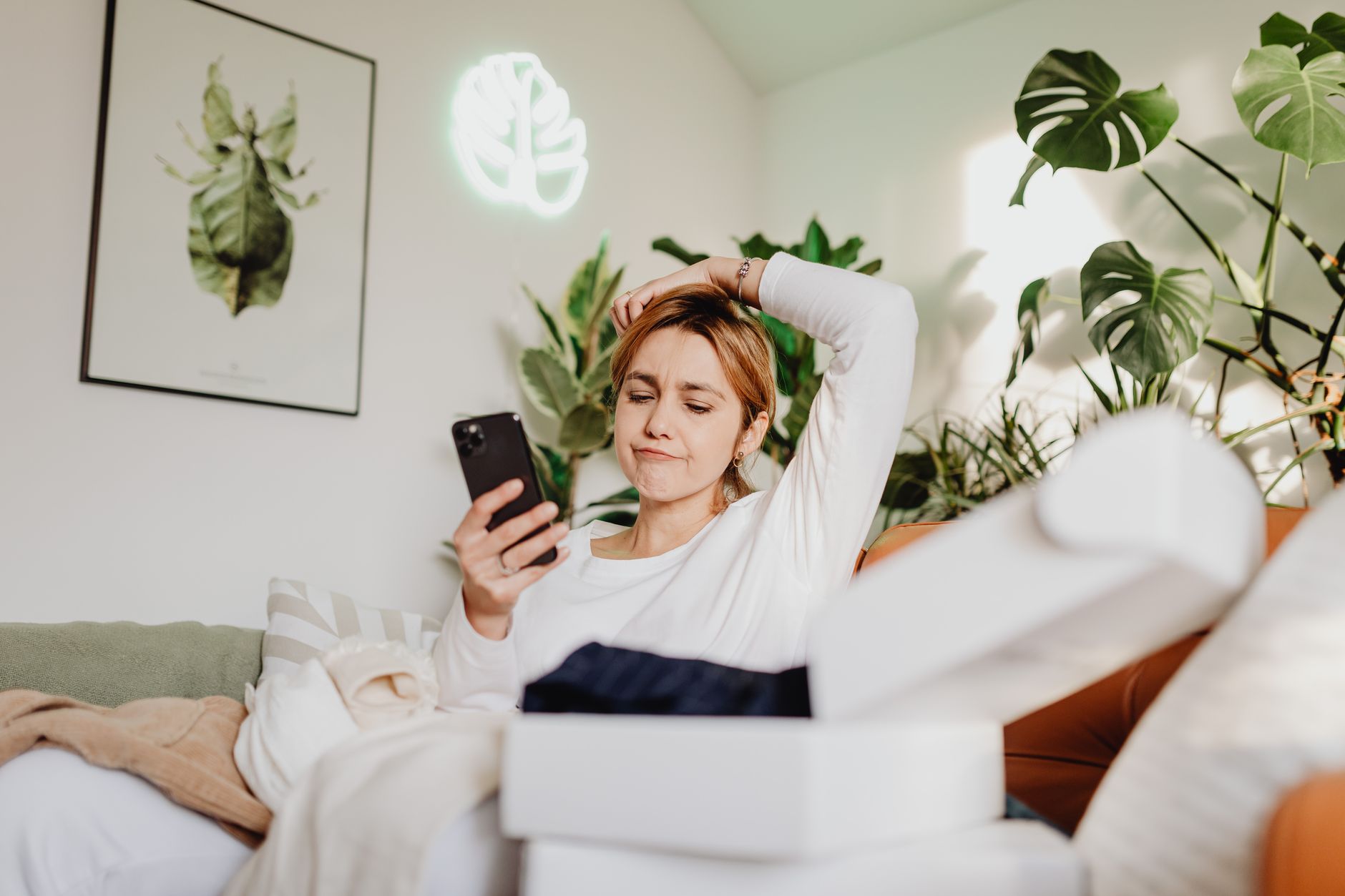 A woman sitting on a couch surrounded by plants, using her phone with a thoughtful expression.
