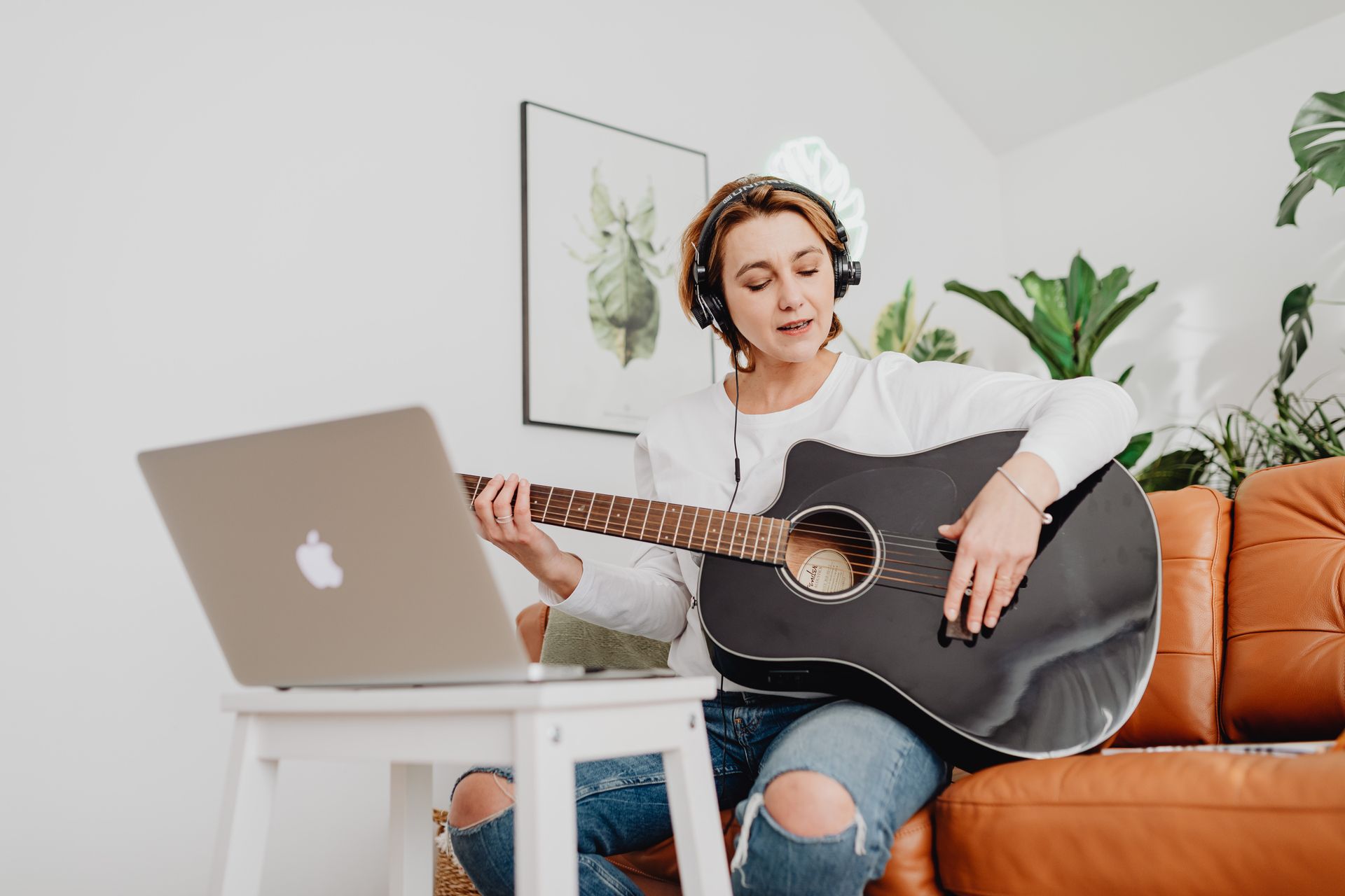 Woman playing guitar with headphones in a cozy living room, using laptop for music tutorials.