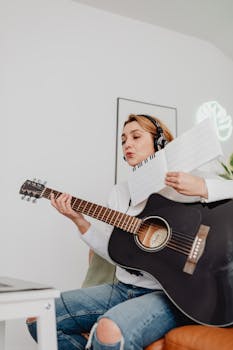 A young woman practices guitar indoors while wearing headphones, studying from sheet music.