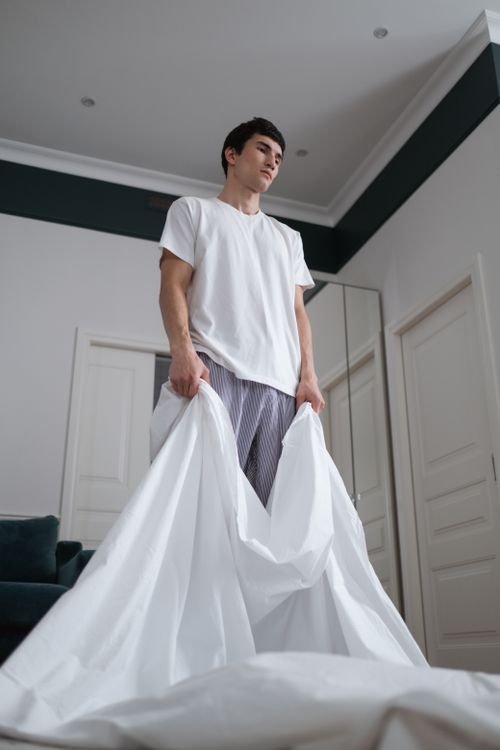 Free Young man in white t-shirt arranging bed sheet in a contemporary bedroom. Stock Photo