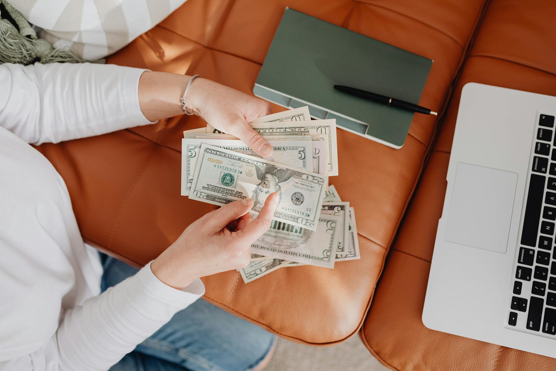 Person holding US dollar bills on brown sofa beside laptop and notebook, representing financial management at home.