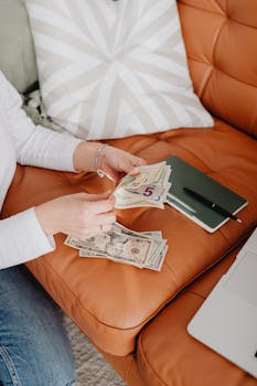A person counts US dollar bills on a leather couch, next to a notebook and laptop.