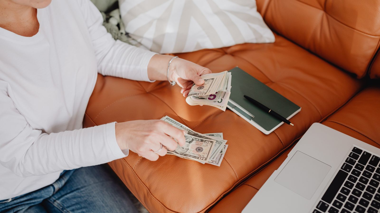 A woman counting cash on a sofa with an open laptop and notebook nearby