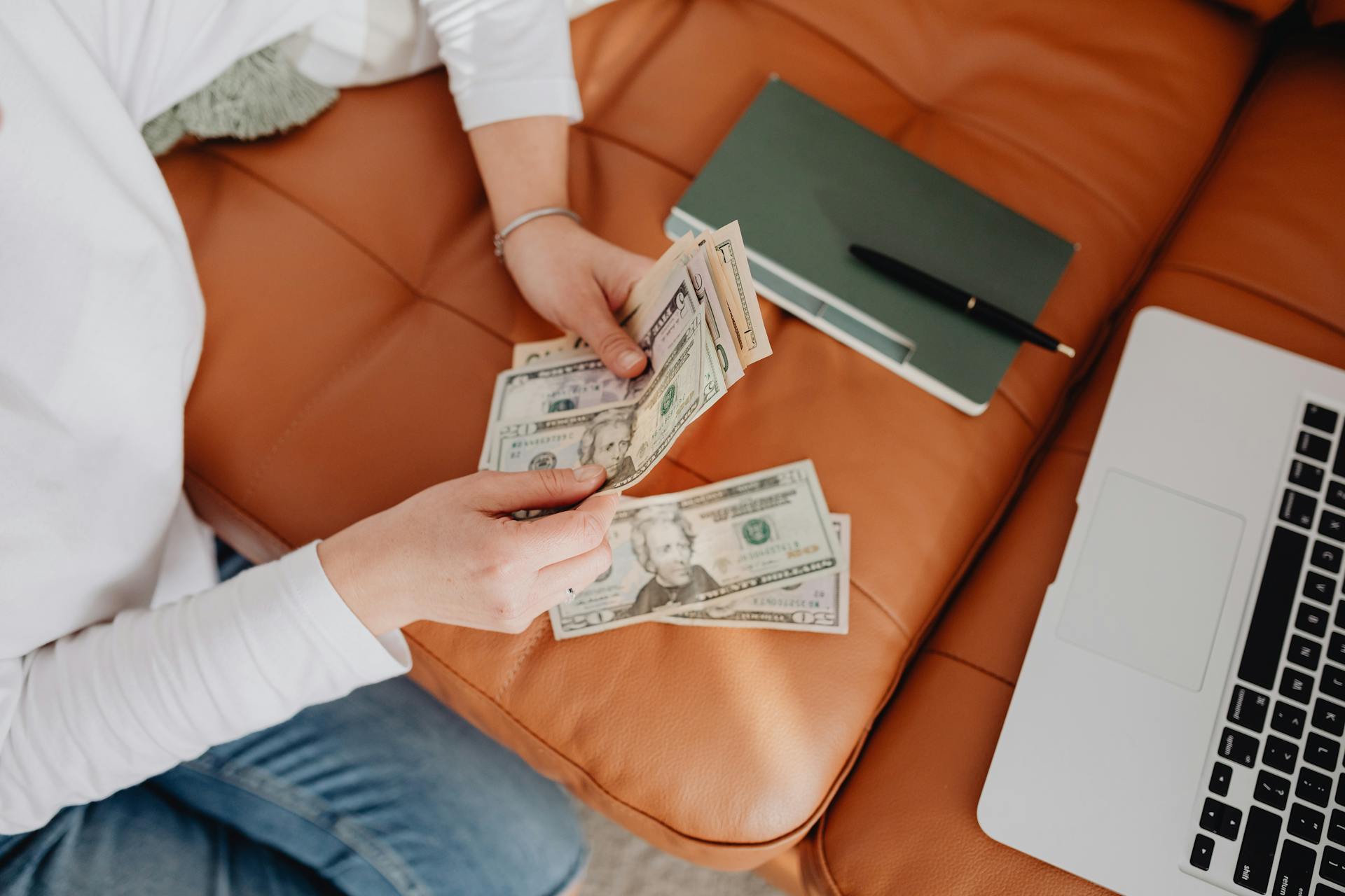 Person counting dollar bills on a sofa next to a notebook and laptop.