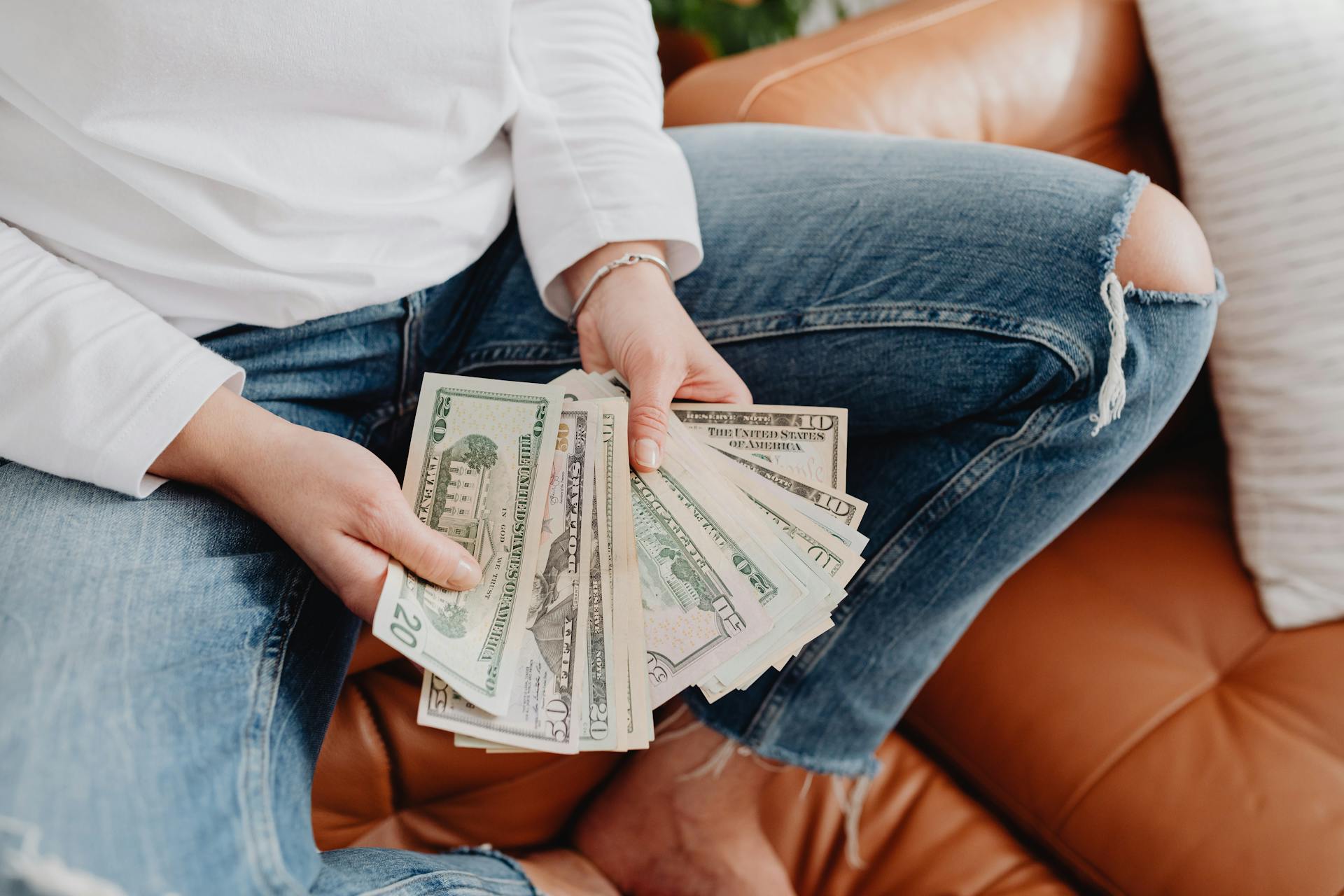 A woman holding and counting US dollar bills while sitting on a leather sofa wearing jeans.