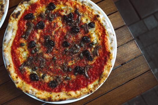 Aerial view of pizza with anchovies, black olives, and tomato sauce on wooden table.