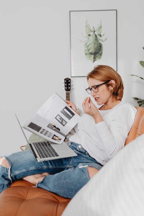 Free Woman in White Long Sleeves Shirt Sitting on a Couch with Her Laptop while Holding a Magazine Stock Photo