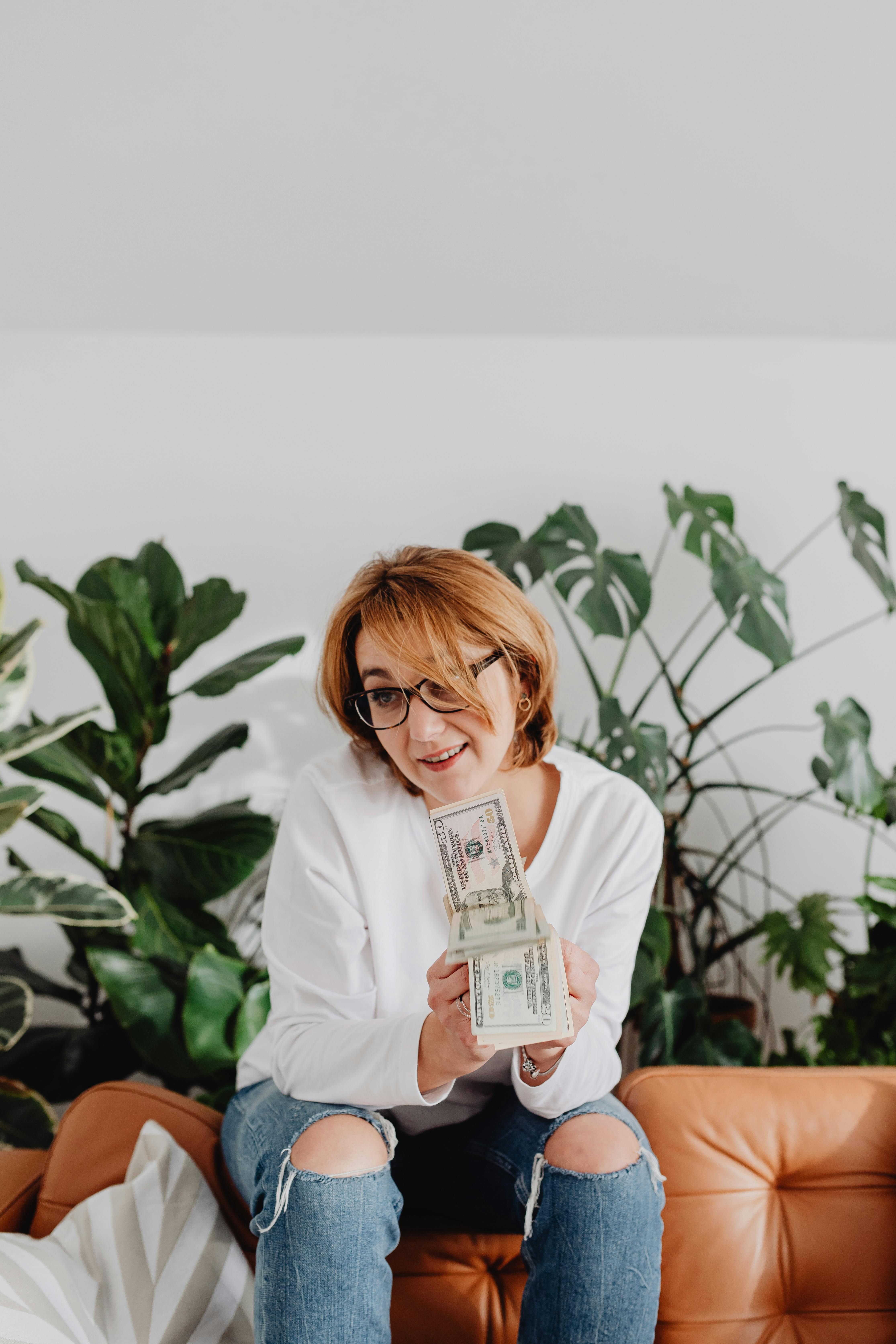 一位微笑的女人穿着白色长袖，拿着纸钱坐在沙发上，旁边有室内植物