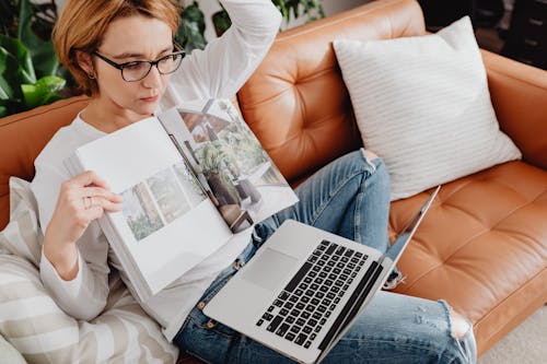 Free Woman in White Long Sleeves Shirt Sitting on a Couch with Her Laptop while Holding a Magazine Stock Photo