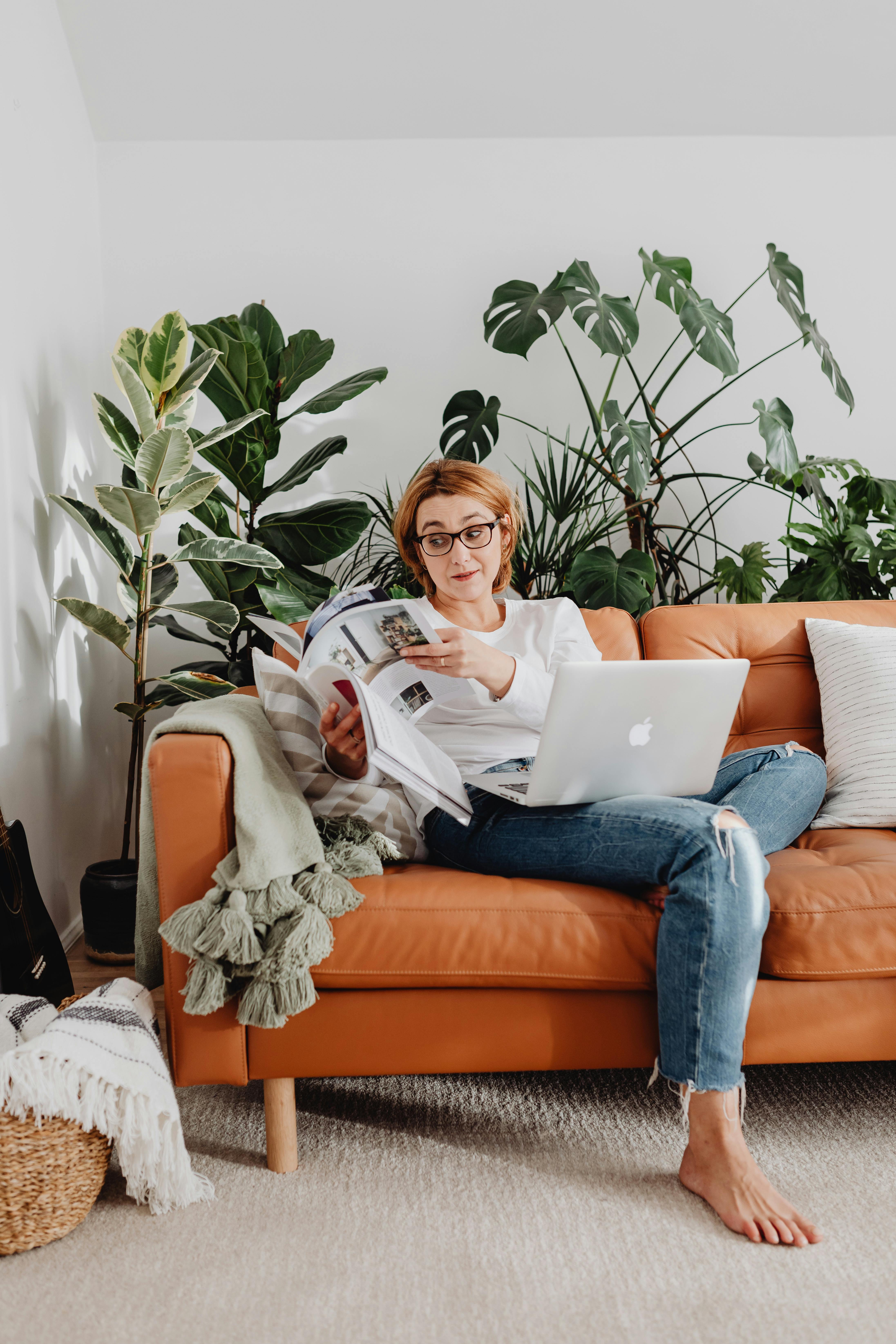 A woman enjoys remote work from home on a cozy couch surrounded by plants.