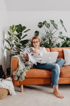 A woman enjoys remote work from home on a cozy couch surrounded by plants.