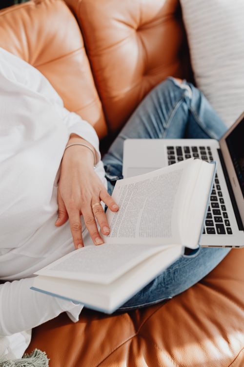 Free Person reading a book while using a laptop on a comfortable leather sofa. Stock Photo