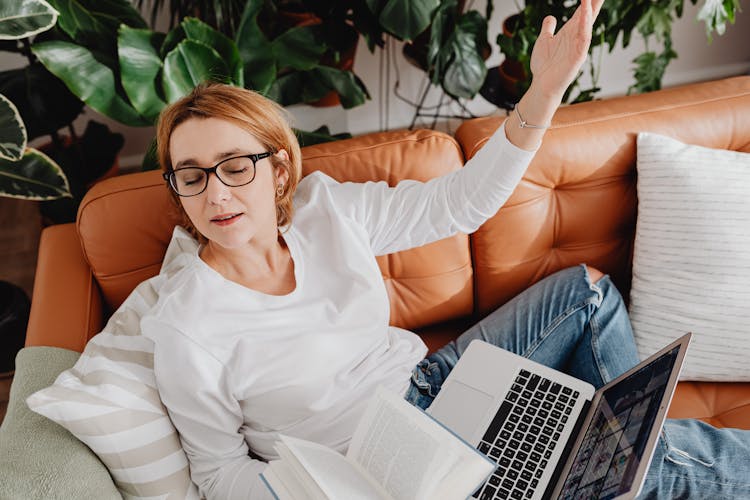 Woman Resting On A Couch While Holding An Open Book And Laptop