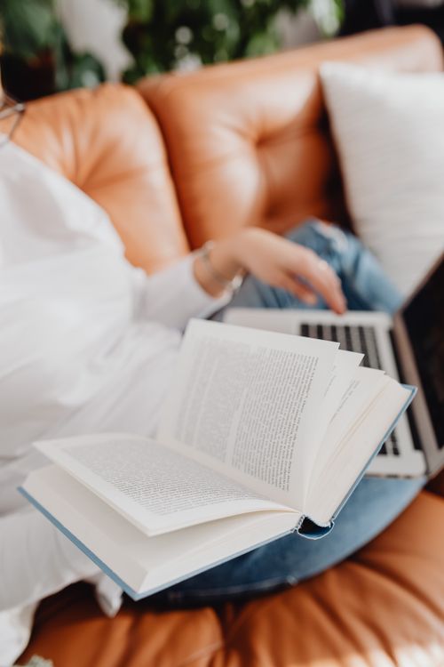 Free Individual relaxing with a book and laptop on a comfortable leather couch. Stock Photo