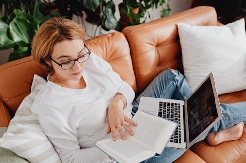 Woman in White Long Sleeves Sitting on a Couch while Reading a Book with Laptop Near Her