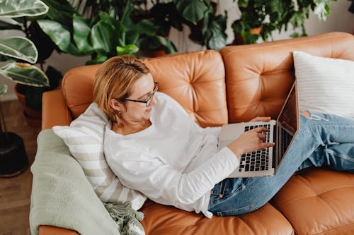 Free Woman in White Long Sleeves Shirt Typing on Her Laptop while Resting on a Couch Stock Photo