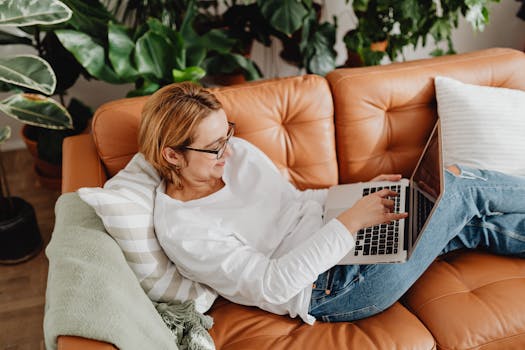 Woman in casual attire using a laptop on a couch surrounded by plants, emphasizing remote work culture.