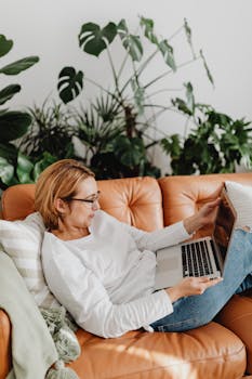 A woman enjoys remote work from home, lounging on a leather couch with plants in the background.