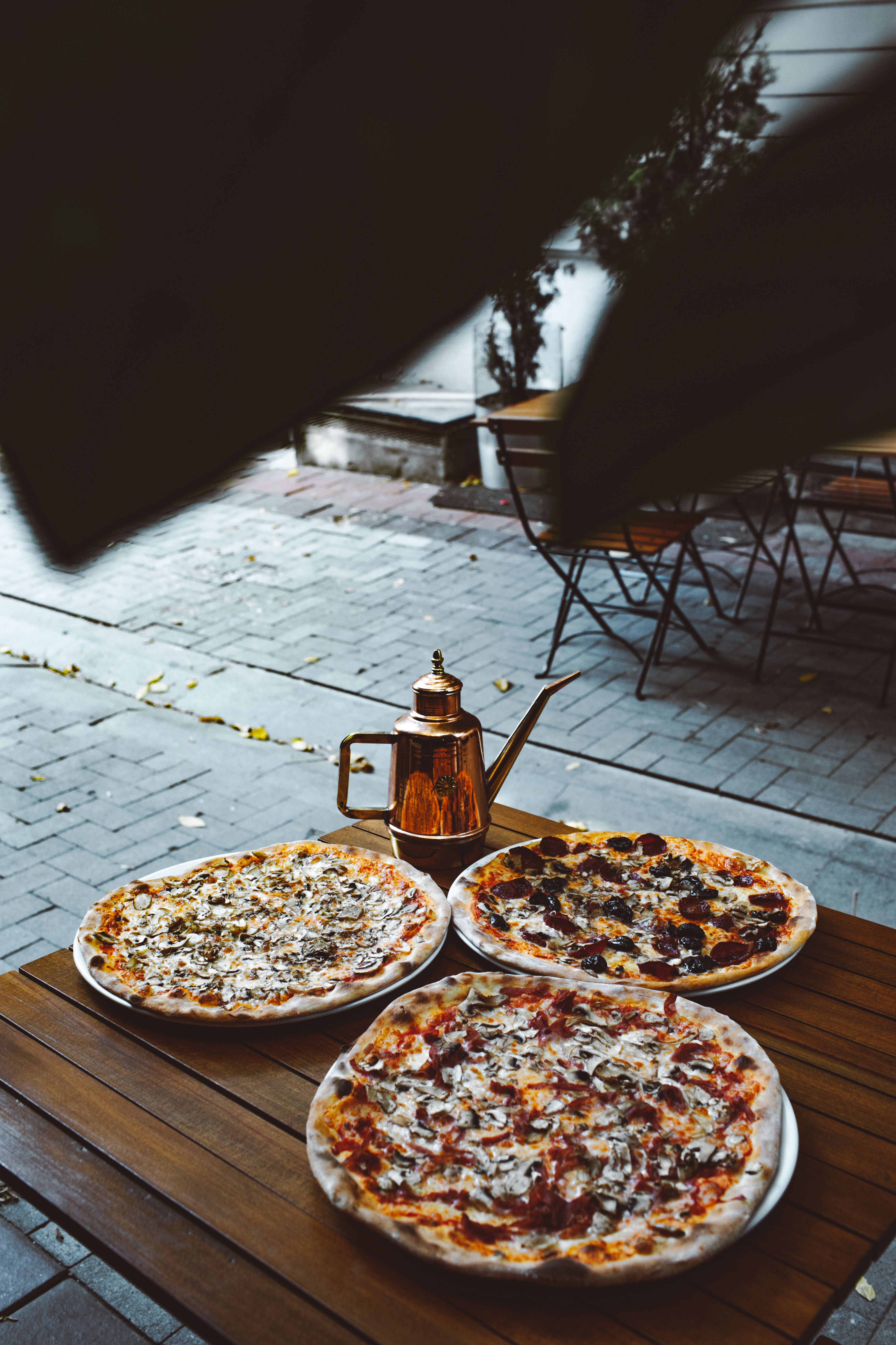 Three delicious pizzas on a wooden table outdoors with a decorative kettle, creating a warm and inviting atmosphere.