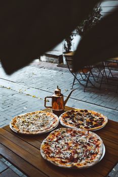Three delicious pizzas on a wooden table outdoors with a decorative kettle, creating a warm and inviting atmosphere.