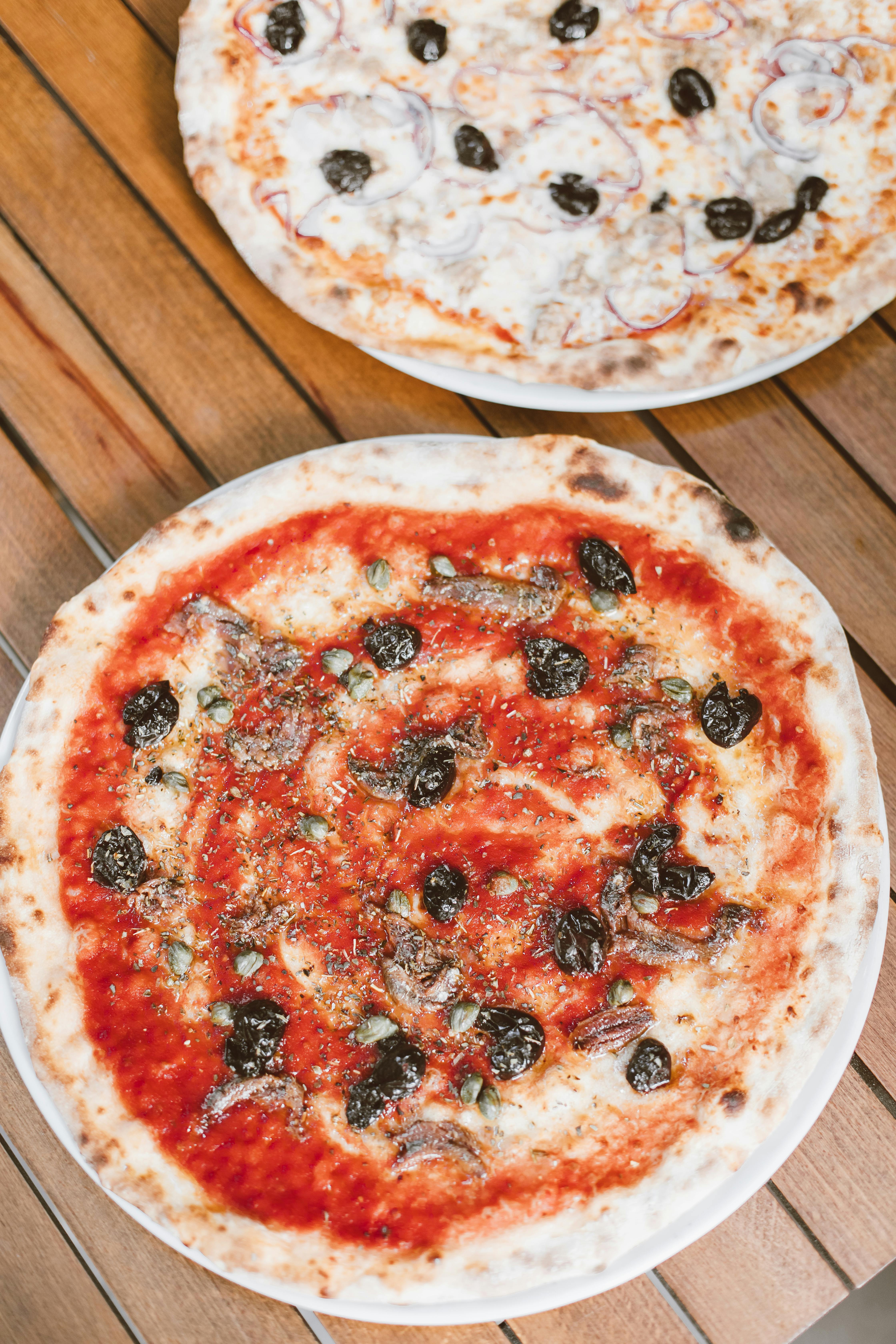 Two Mediterranean-style pizzas with black olives on a rustic wooden table.