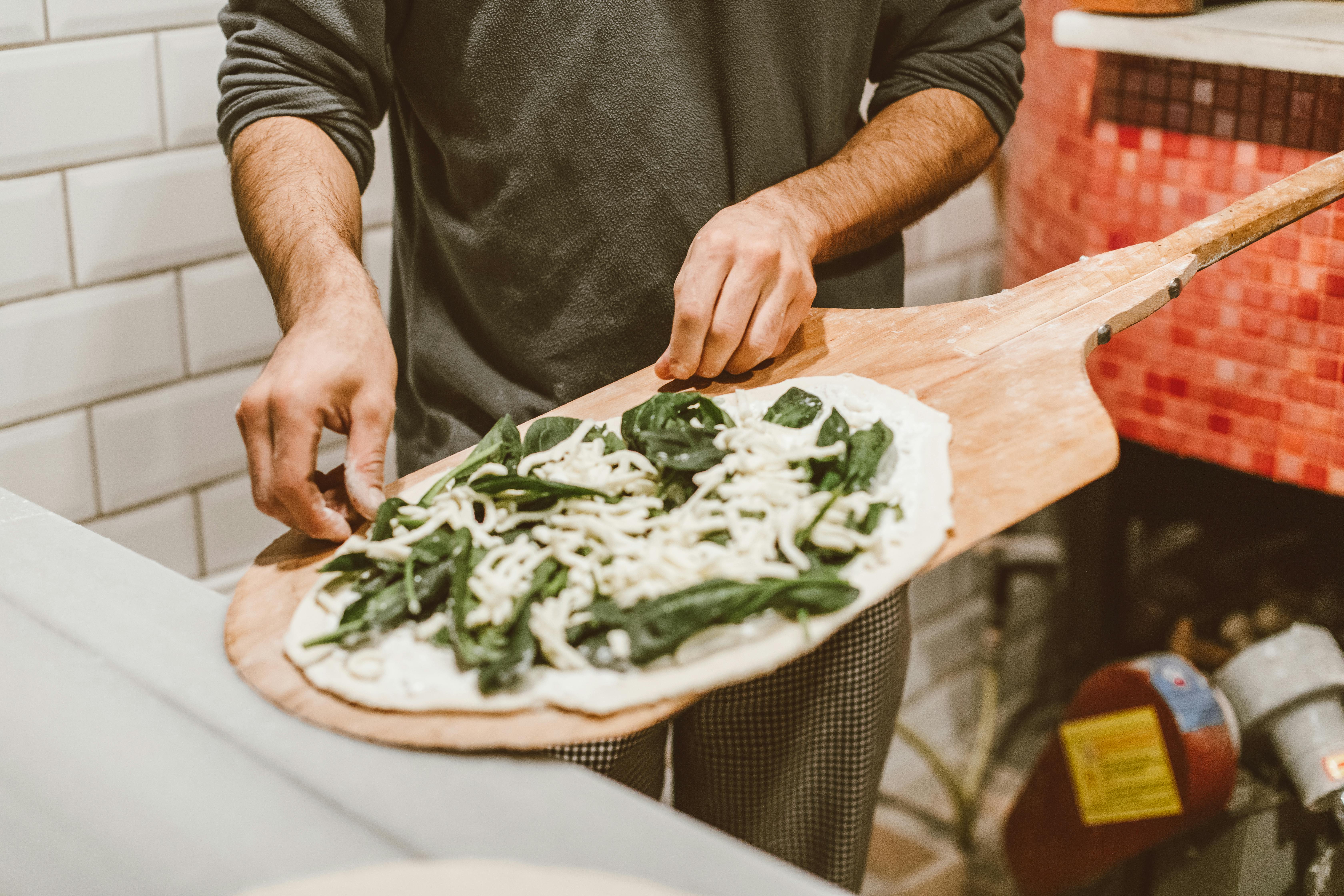 A person crafting a spinach and cheese pizza with a wooden paddle near a traditional rustic oven.