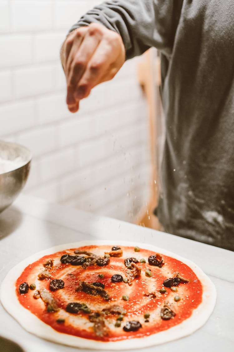 A Hand Sprinkling Cheese On A Pizza Dough With Red Sauce