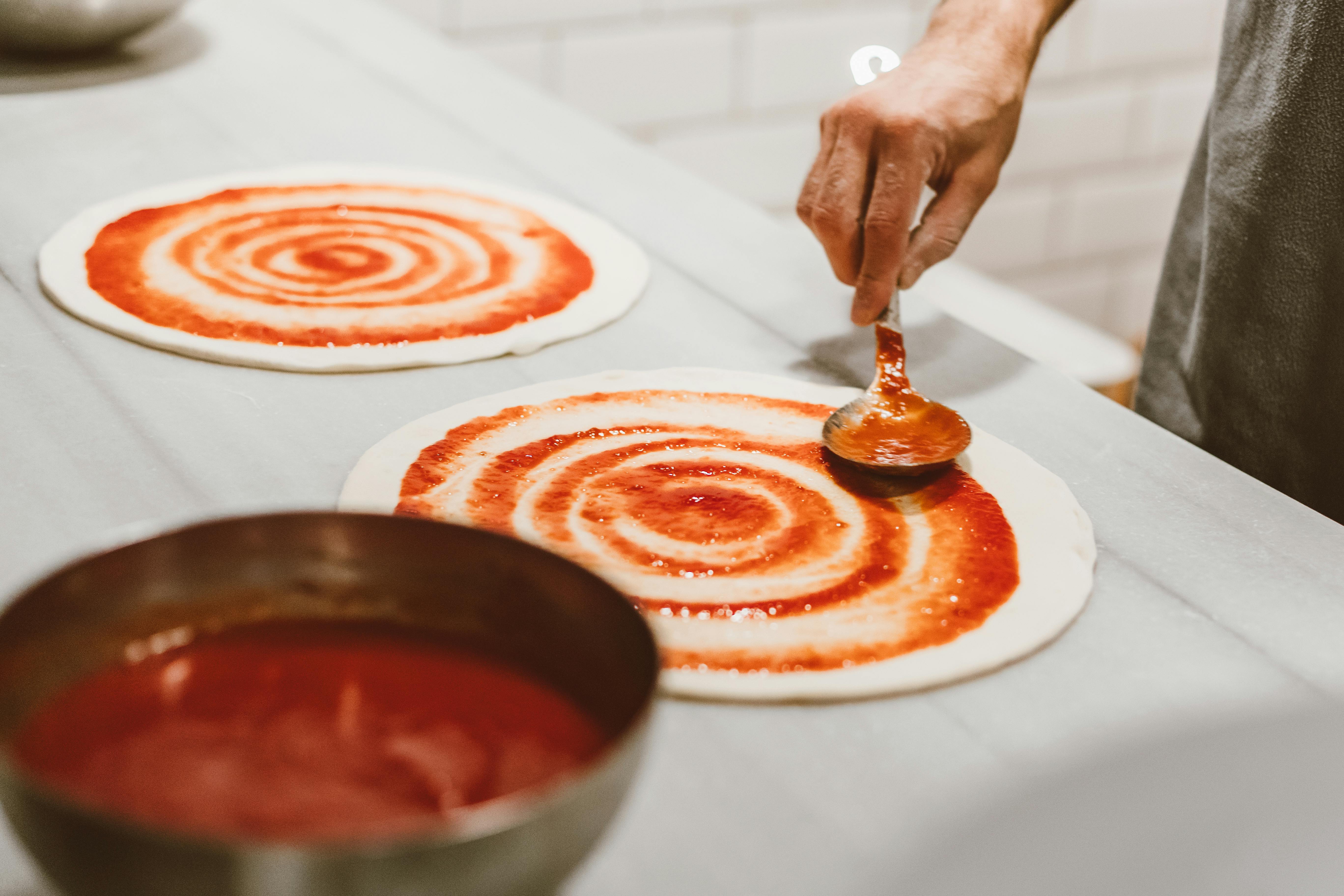 Hand spreading tomato sauce on pizza dough in an indoor setting.