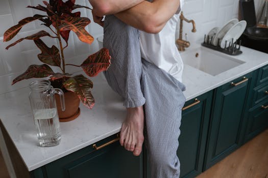 Person sitting on kitchen counter with house plant and glass pitcher nearby.