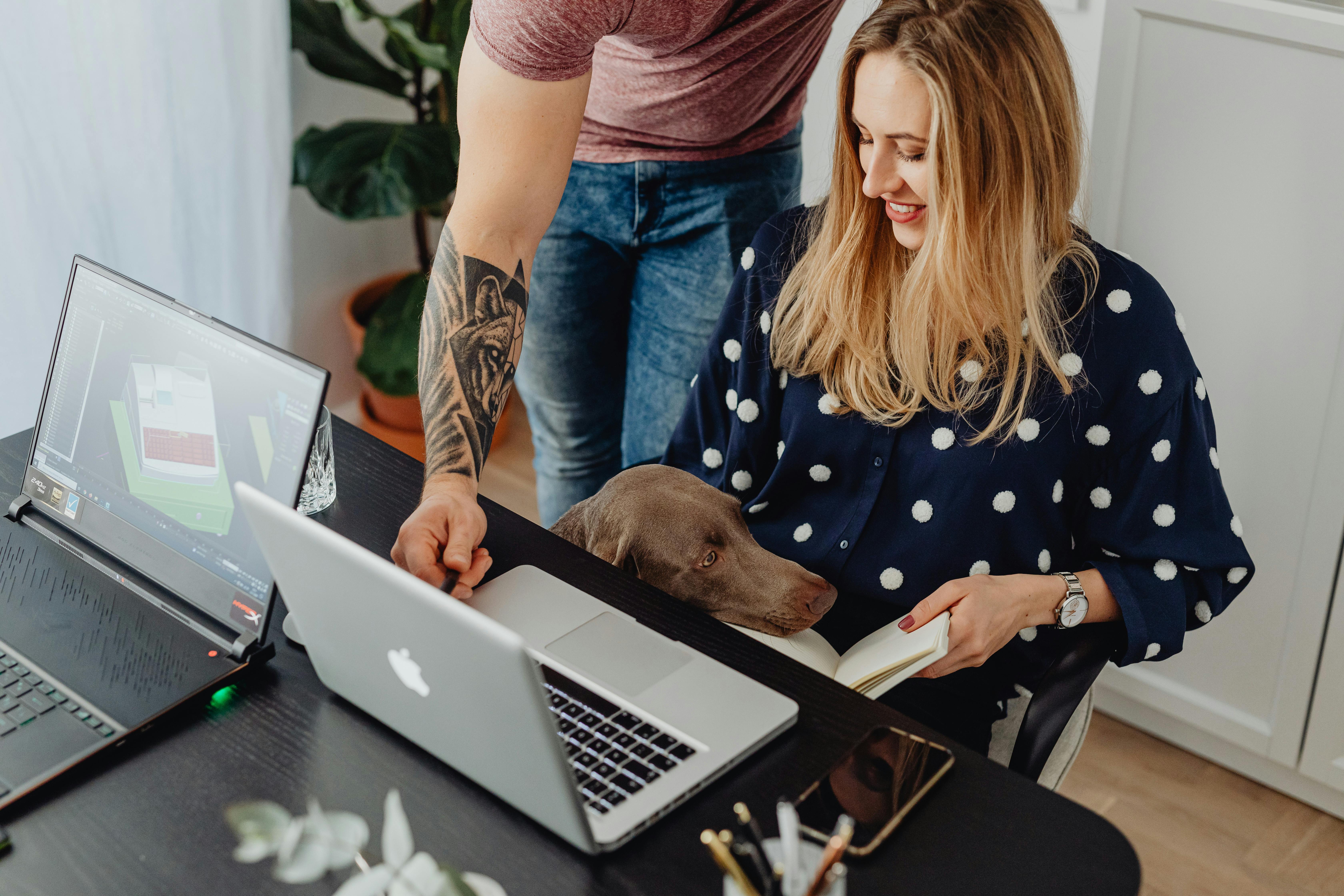 A woman multitasking at home with a laptop, book, and her dog for company.