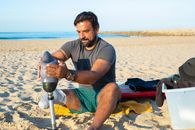 Tanned man in casual clothes putting on artificial limb leg while spending time on seashore