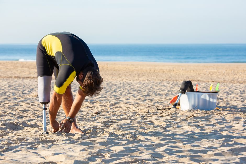 Side view of male in wetsuit and with artificial limb bending forward while stretching on sandy coast in sunlight