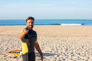 Happy male in diving suit looking and pointing at camera while standing on beach in bright sunny day