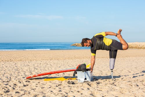 Side view of male in wetsuit and with artificial limb stretching leg holding on box while preparing for surfing