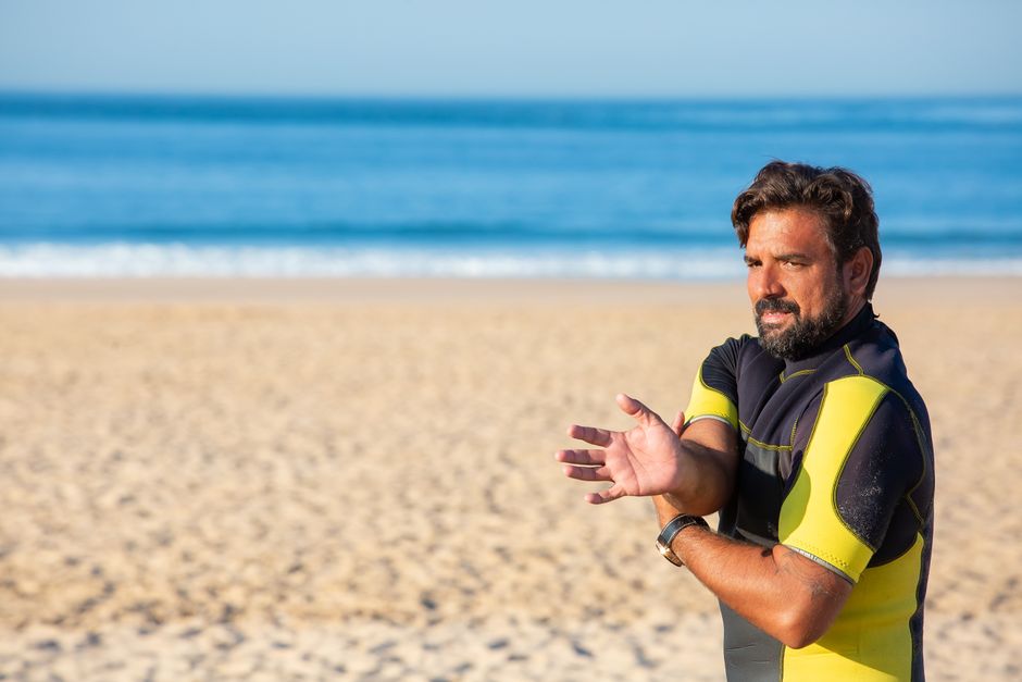 Tanned male in wetsuit doing exercise for stretching hand while standing on sandy beach