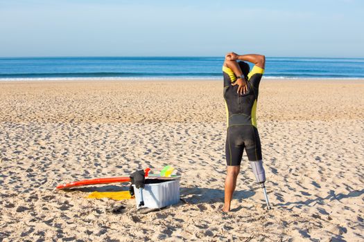 Back view full body male amputee surfer in wetsuit stretching body on sandy beach before surfing
