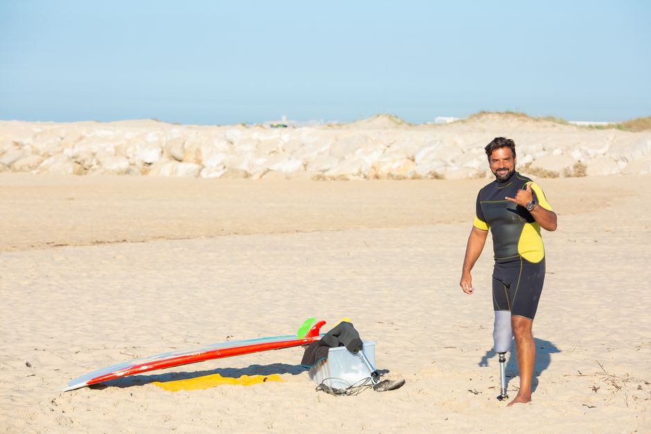 Full body content ethnic amputee male surfer wearing wetsuit standing on sandy beach and showing shaka sign while looking at camera with smile