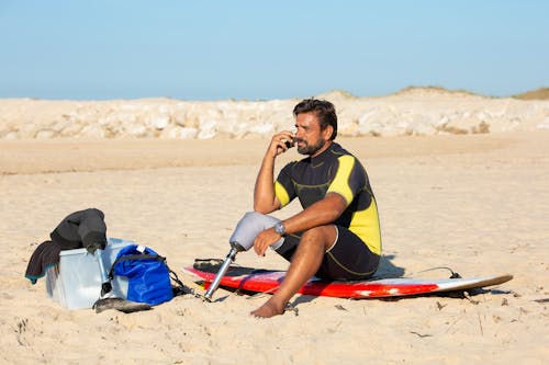 Full length tanned bearded male surfer with leg prosthesis sitting on sandy beach on surfboard and having conversation on mobile phone